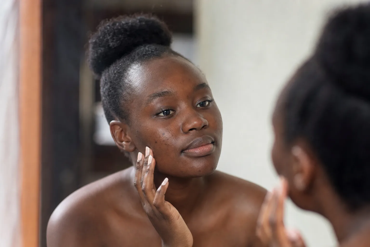 Young Black African woman with deep brown skin and a few pimples on her cheek looking calmly into a mirror in soft daylight