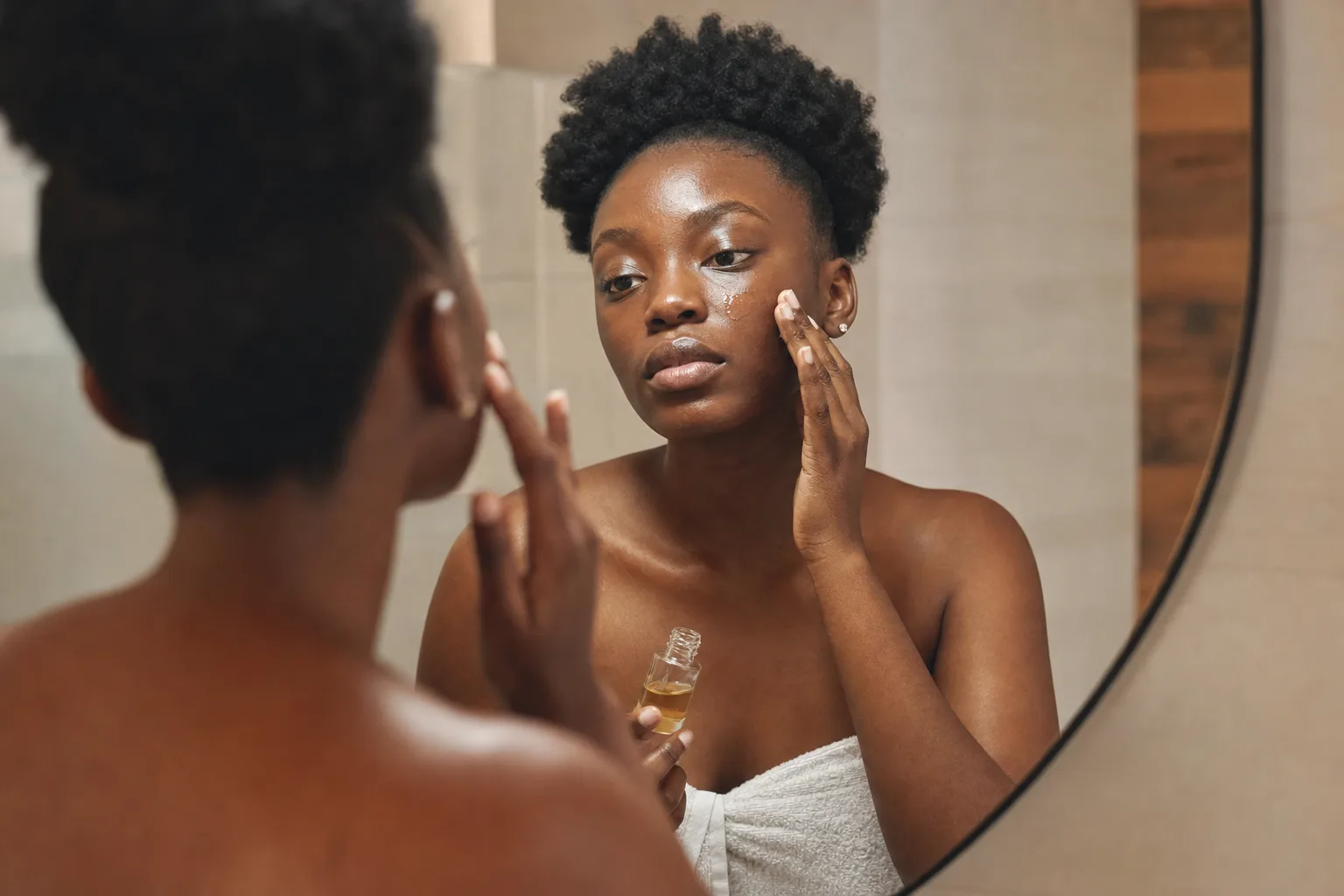 Black African woman with deep brown skin smoothing a treatment serum onto her cheek in front of a mirror in warm bathroom light