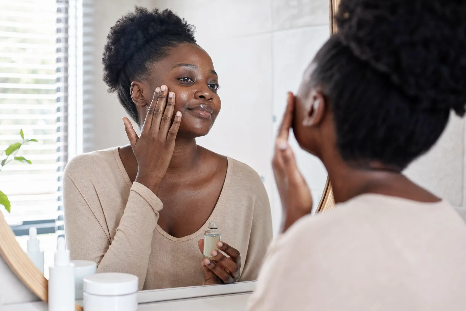 Black African woman applying skincare in front of a mirror with a few products arranged on the counter, bright natural light